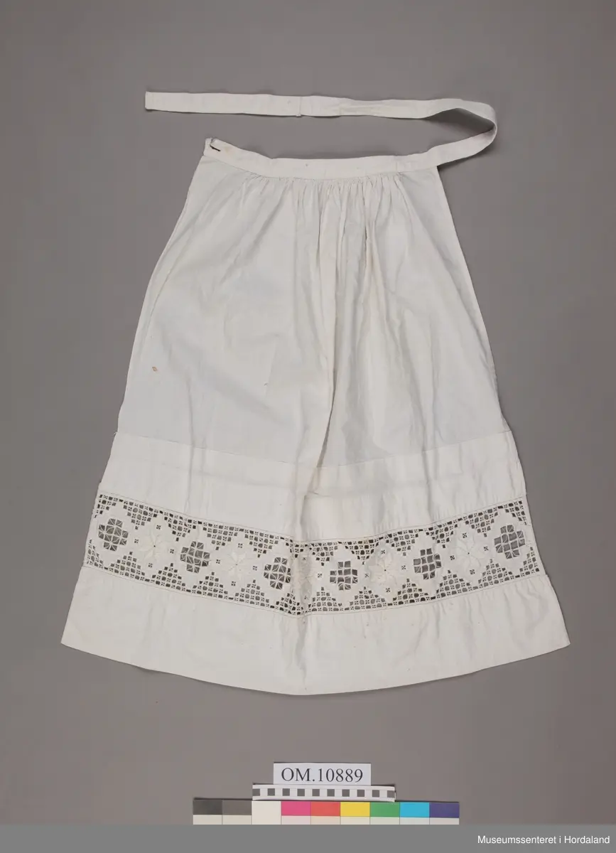 Form: bunadsforkle med linning og band i livet, knappa bak, innfeld broderi nedst på forkle
