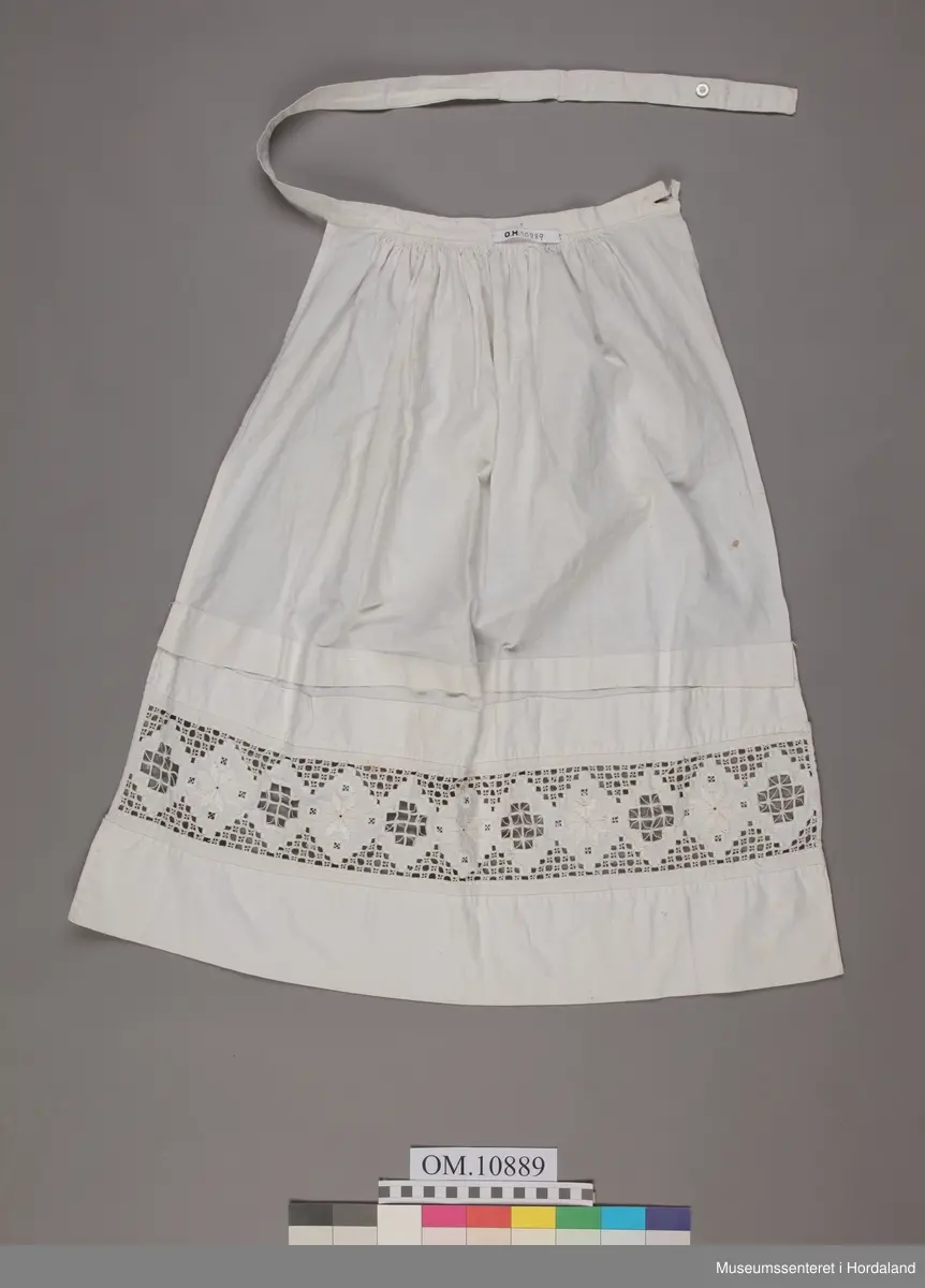 Form: bunadsforkle med linning og band i livet, knappa bak, innfeld broderi nedst på forkle
