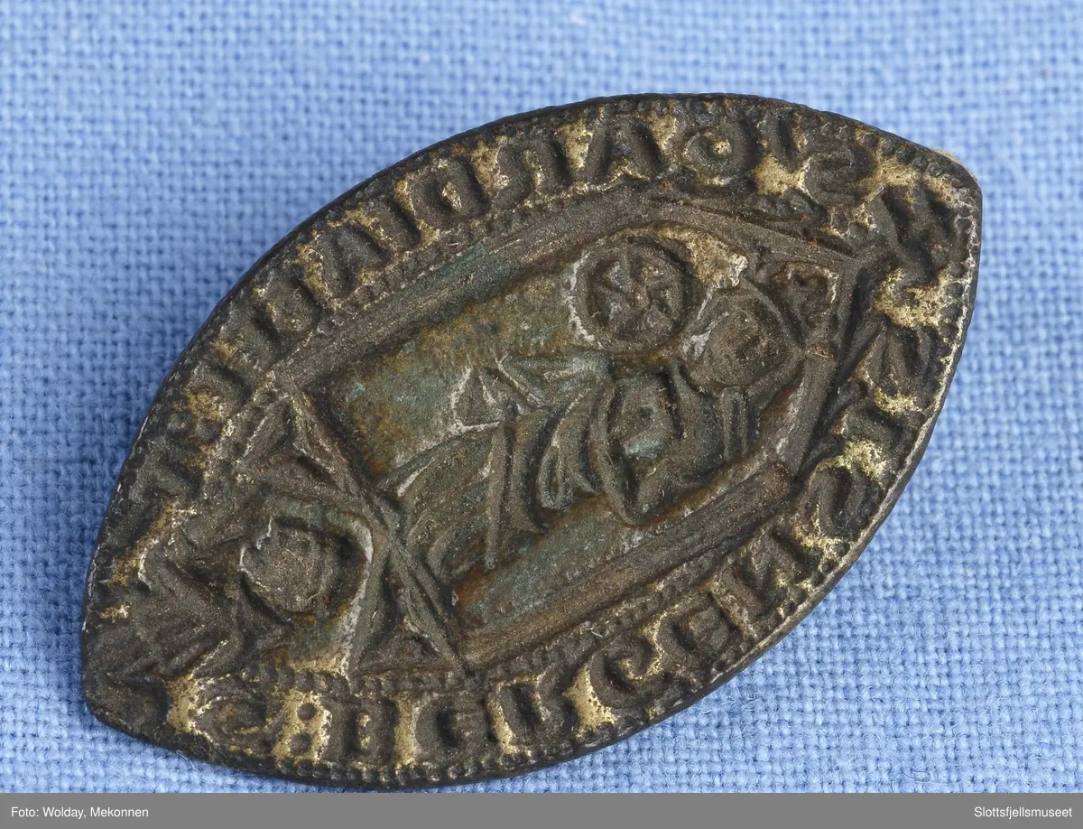 Spissoval form og innenfor omskrift står en kronet skikkelse som holder et hjul eller lignende. Bedende person nederst, muligens en munk.