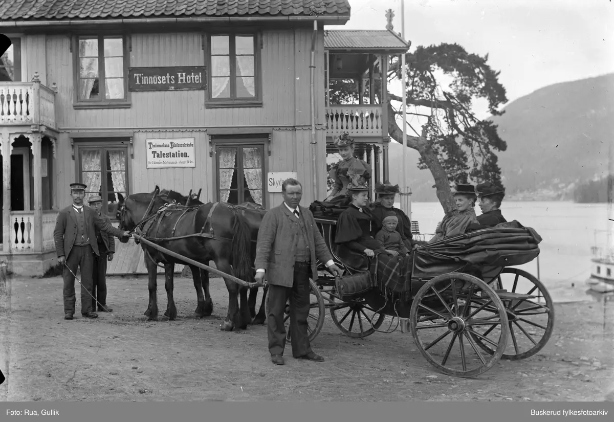 Hest og vogn med passasjerer står på skysstasjonene utenfor Tinnosets Hotel. Midt i bildet ser vi hotelleier Christensen og ved hestene står vognmann Ole Bogen med sønn som kjørte skyssen fra Kongsberg. På hotellets vegg henger en plakat med teksten "Thelemarkens Telefonselskaps Talestation".
