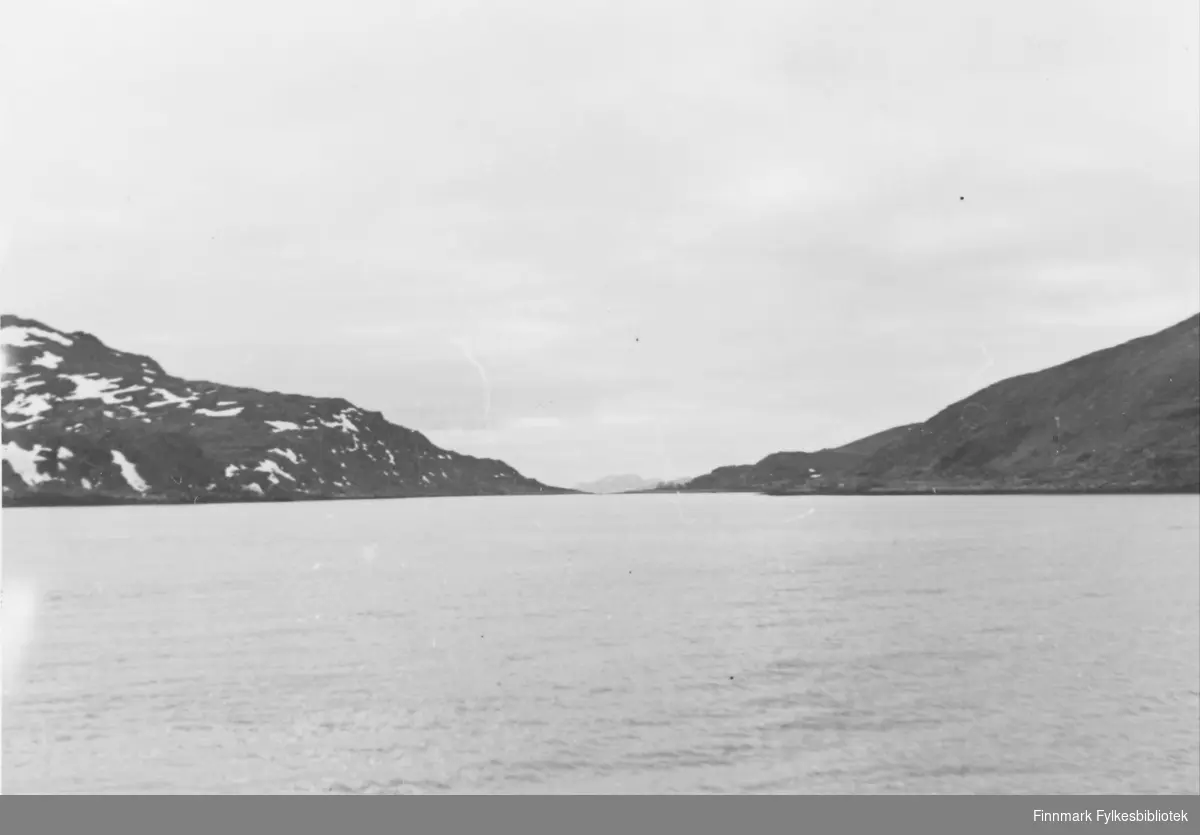 Naturbilde. Tatt utover fjorden ved Havøysund. Slakke fjellskråninger strekker seg ned fra hver sin kant og møtes nesten i vannkanten i bakgrunnen av bildet. Sjøen ligger rolig, det er overskyet.