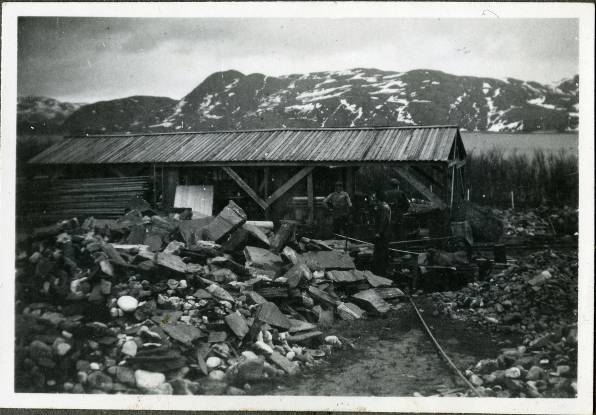 Tre arbeidende menn står foran et bygg som er fullt av redskaper og byggemateriale. Rundt om kring på bakken kan man se skifer, og i bakgrunnen ser man havet og fjell. Mennene er kledd i arbeidsklær.