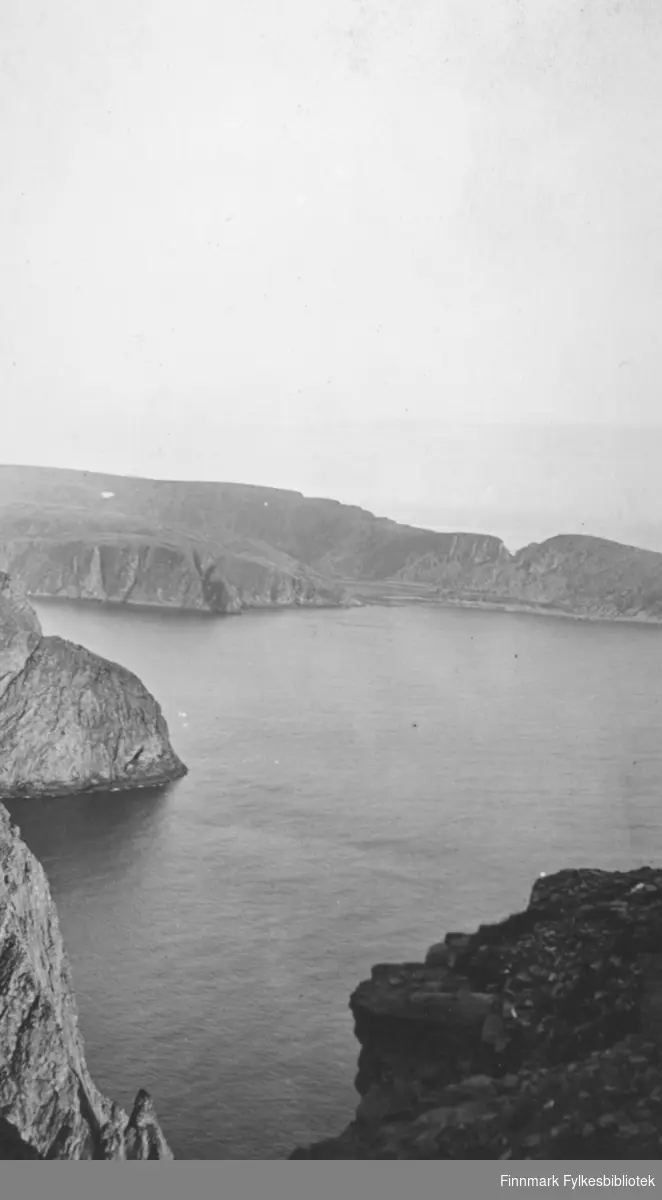 Bakerst på bildet er deler av Knivskjellodden fotografert fra Nordkapp-platået. Havområdet er Knivskjellbukta som ligger blikkstille i solskinnet. Neset midt på bildet til venstre er fjellet Lille Kappa.