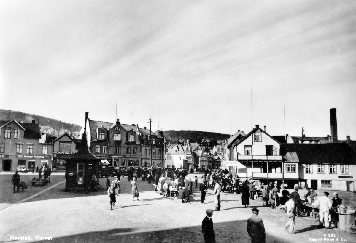 Publikum og torghandlere på torget i Harstad. Kiosk til venstre og oppstilte drosjer i bakgrunnen.