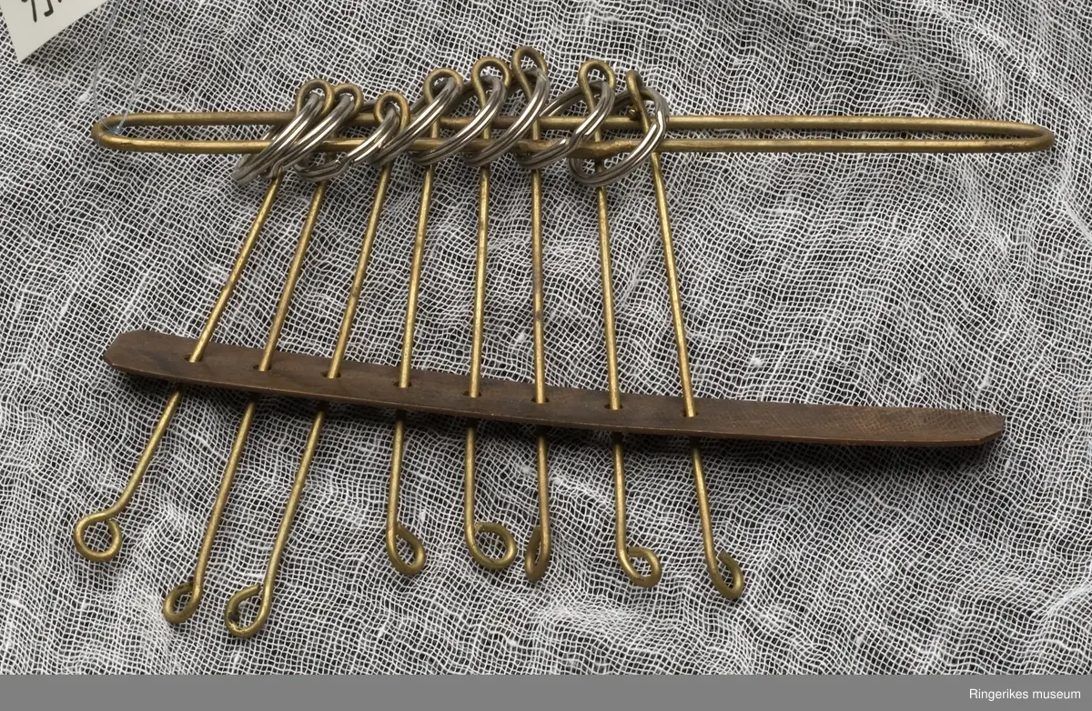 Bøyd og formet messing tråd, loddet i skjøten. Åtte ringer tredd inn på buen, til hver ring festet en rett messingtråd med bøy i begge ender. I enden av messingtråden er tredd på en kobbberplate med tilsvarende  åtte hull. De åtte messingtrådene i rammen er bevegelige. Leken elelr apparatet har i sin sammensetning en rektanuglær form. Ukjent funksjon og bruk.