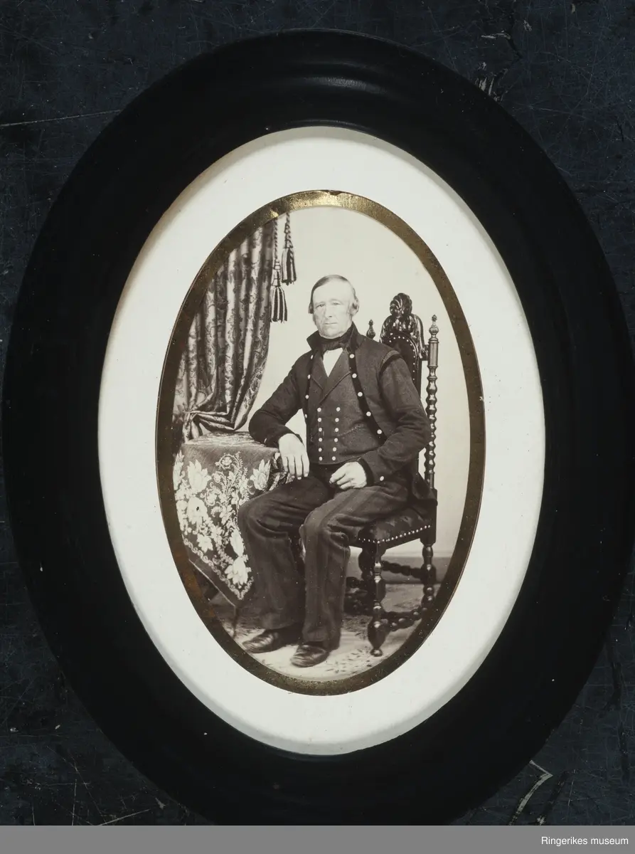 Svart/hvitt fotografi i svart oval ramme. Passpartur med gullkant inn mot fotografiet. Påskrift bakpå rammen.