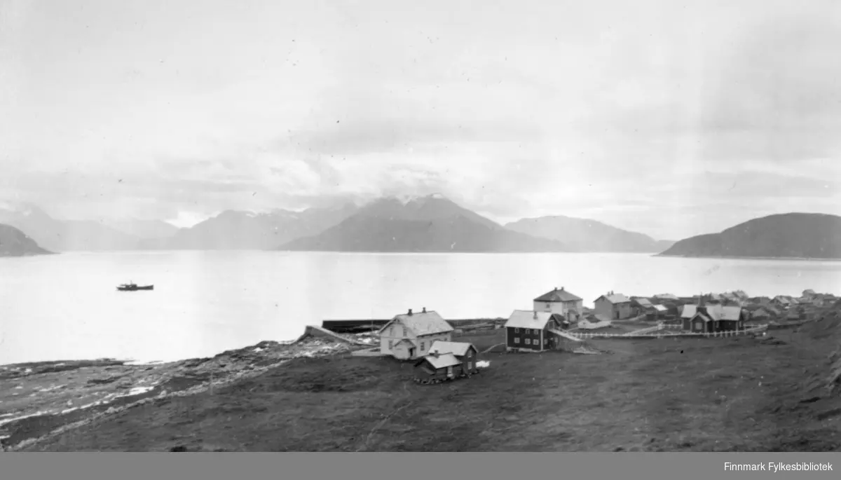 Loppa øy i Loppa kommune. Bebyggelsen ligger i strandkanten. Et ganske stort hus nærmest med fjøs og et ganske lite bygg. Ellers ligger husene i en klynge ved sjøen. Et stort jorde ligger foran bebyggelsen og en båt er ute på den blikkstille fjorden. Noen snøflekker ligger på bakken til venstre på bildet. På andre siden av fjorden ses fjell, noen av dem med snø på toppene. Skyet, men bra vær en sen vårdag.