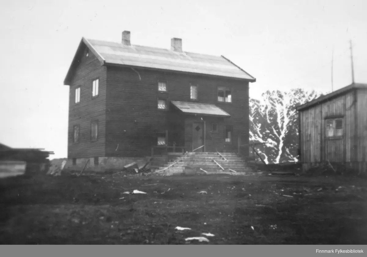 Et stort hus med en bred trapp. Et vindfang står midt på langveggen. Huset er malt med en mørk farge og har vinduer av ulik størrelse på langveggen, og to vinduer på røstveggen. To skorsteiner står på det pappkledte taket. En lav grunnmur har to små vinduer og til venstre for huset ligger en stabel med ved/materialer. Bakken foran huset består stort sett av jord/grus. Til høyre på bildet ses deler av røstveggen på et lite bygg. Det har stående, umalt panel og et sprossevindu som vises på bildet. Et stort fjell ses mellom bygningene. Det er endel snøflekker på det.