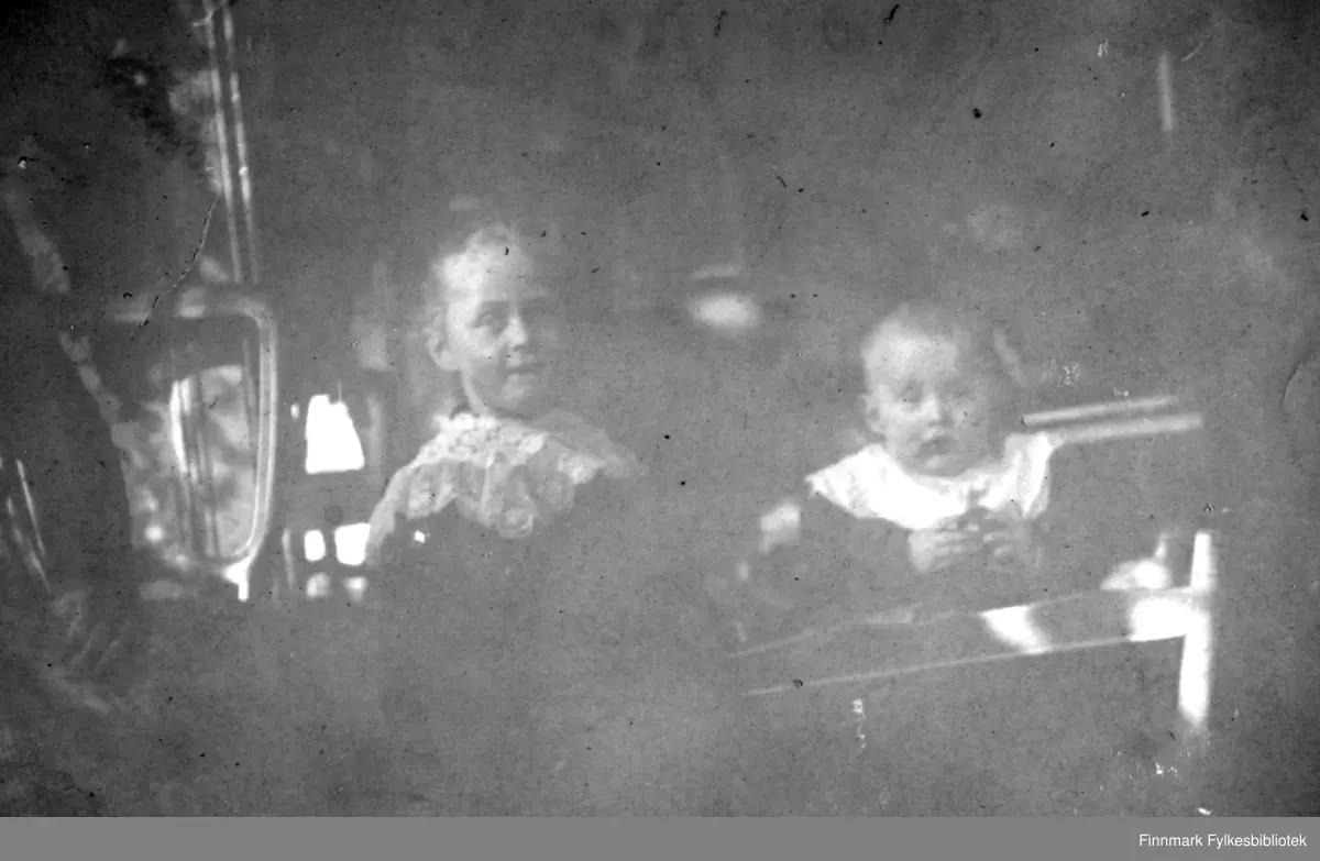 Tre personer fotografert i et rom. Til venstre sitter en dame i rutet skjørt som hviler armene sine fanget. I midten er en ung jente i mørk kjole. Hun har et hvitt, lite sjal rundt halsen. Til høyre sitter et spedbarn. Barnet har mørk påkledning med en hvit smekk rundt halsen. Et lite bord/brett står foran barnet. I bakgrunnen ses ryggen på en stol og en lampe stående på et bord.