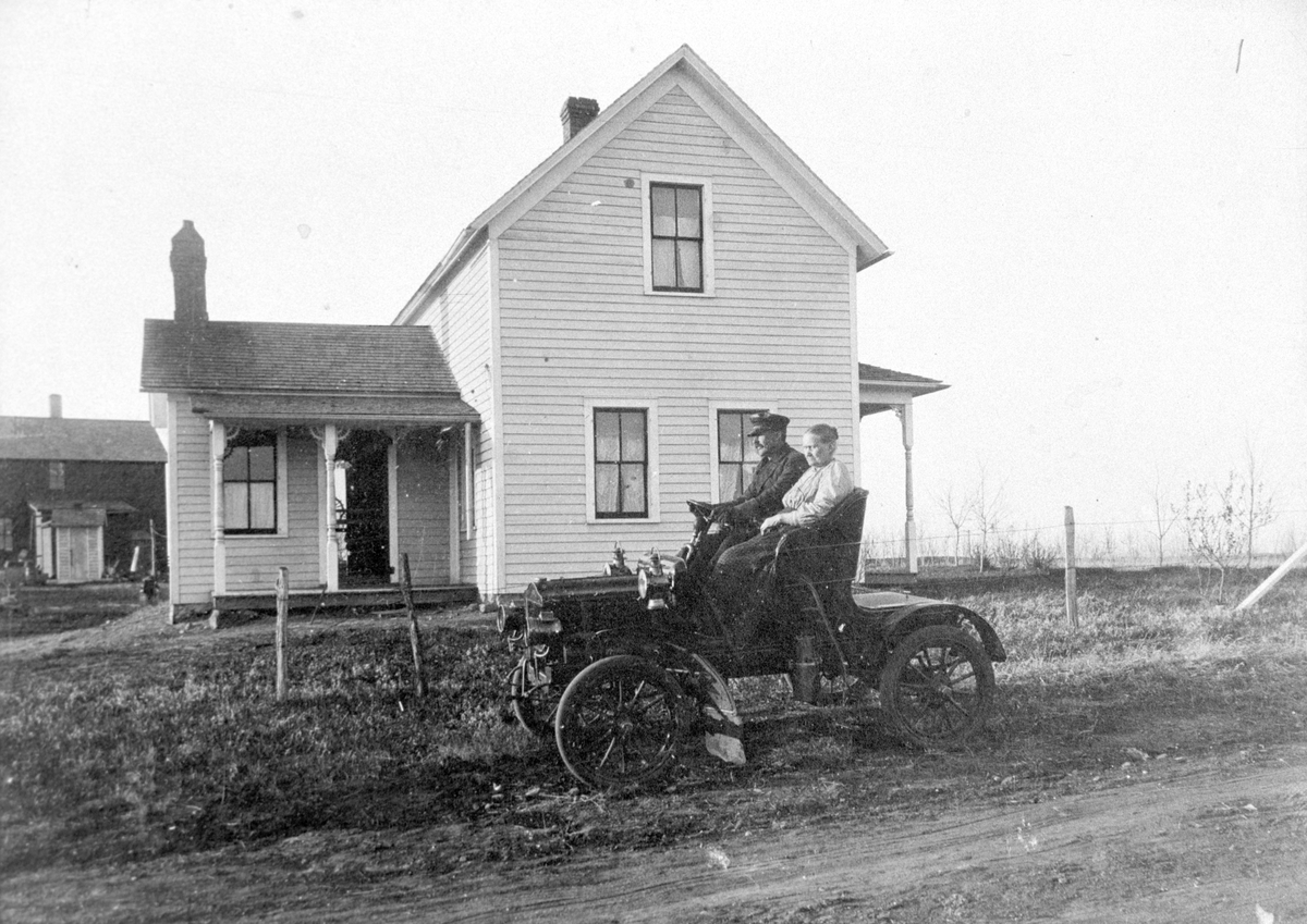 Amerikabilder, Per Mæhlum, Vallset.
Bil med to passasjerer, bolighus i bakgrunnen, Eastedge, Nord-Dakota, USA. Mannens navn er Remmen, og kvinnen ved siden av er hans mor. Disse to var fra Stange. 
Fotograf er Per Mæhlum, ca 1910.