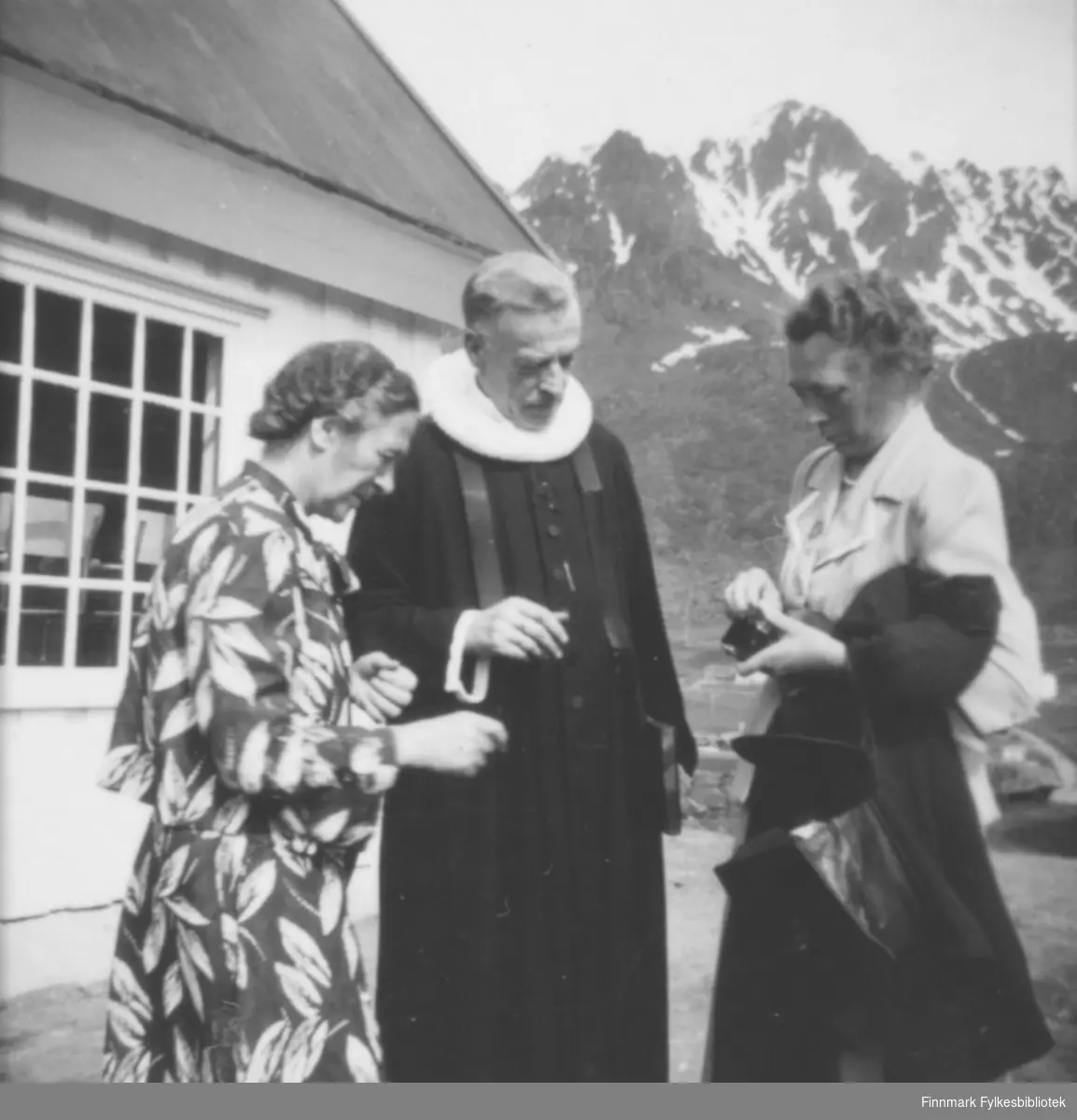 Fylkesprost (senere biskop) Alf Wiig iført sort kappe og krave står mellom to damer ved innvielsen av Bergsfjord kapell eller kirke i Loppa 12.7.1951. Alf Wiig og sogneprest Oterholm arbeidet med reisinga av kirka i Bergsfjord. Alf Wiig holder en bok i venstre hånd. Damen til venstre på bildet har en mønstret kjole. Hun til høyre har en lys overdel og en mørk kåpe hengende over armen. Hun har en stor mørk håndveske som er åpen og noe lite og sort i hendene. Det hvite kirkebygget står rett bak dem og vinduet har sprosser. Gjennom vinduet ses flere stolrygger og taket er pappkledt. En plen ses bak dem og bakerst på bildet ses et stort fjell med endel snøflekker på. Mer informasjon om kirkene i Loppa i boken "Loppas historie: Bygdebok for Loppa kommune".