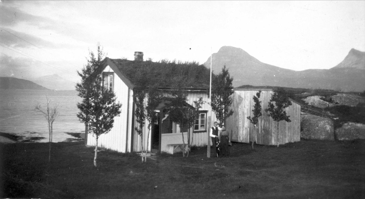 To personer foran en liten stue og et uthus. Sjø og fjell i bakgrunnen.