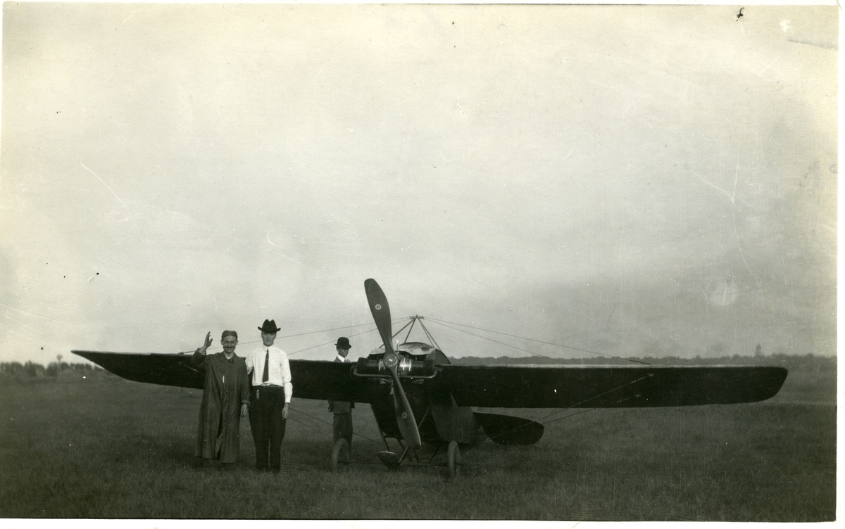 To menn avbildet foran et fly. En tredje mann avbildet bak flyets vinge. To av mennene er iført dress og hatt mens den tredje er iført en form for kappe. Dette er et postkort.