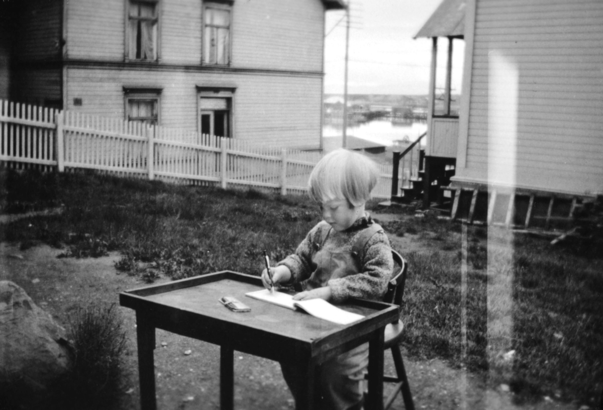 Tor Hauge som sitter med tegnesaker ved et bord, i bakgården til Sellstrømhuset. Huset på andre siden av gjerdet er Jungegården