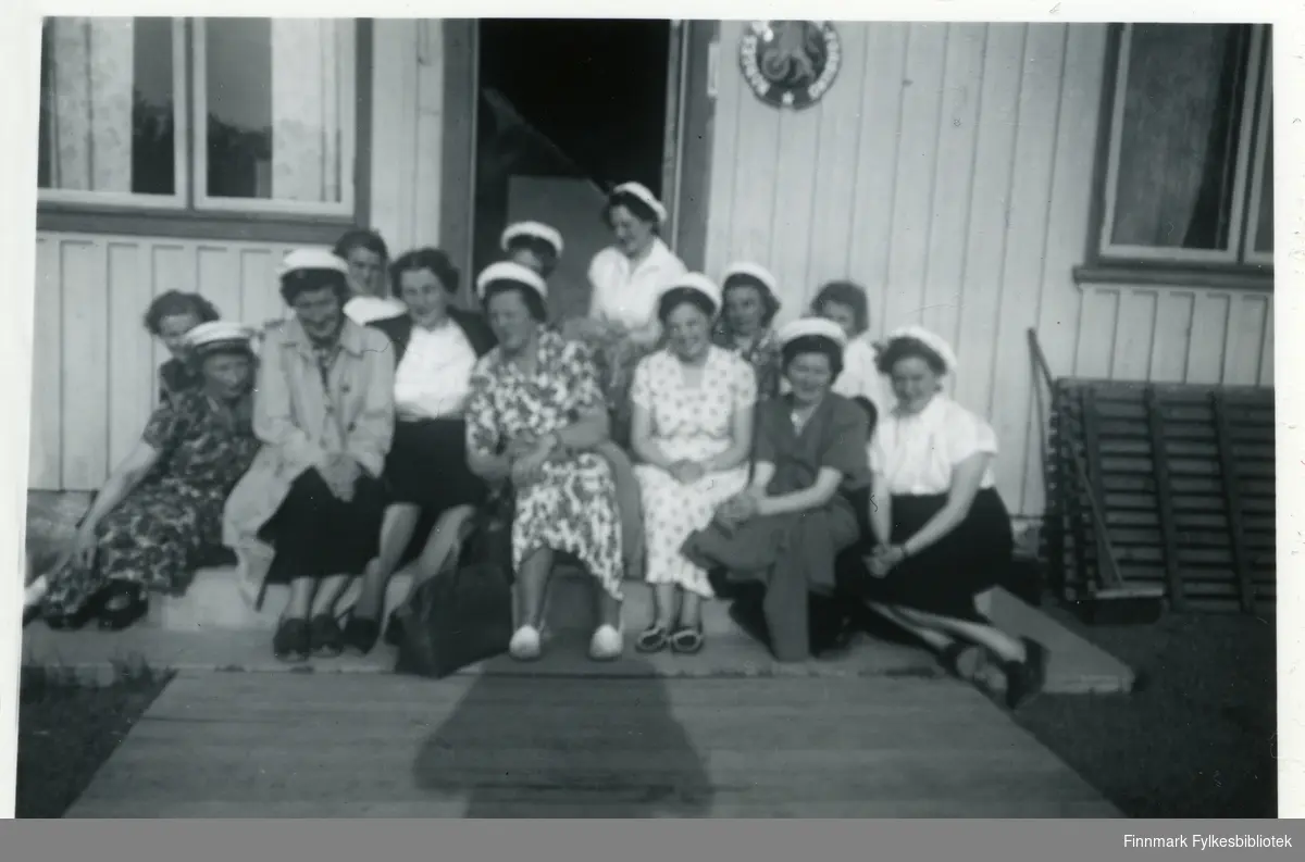 Medlemmer av Vadsø damekor sitter på en trapp og poserer for fotografen utenfor Leif Johnsens Gjestgiveri i Rustefjelbma på en turne-reise. De fleste har på seg hvite hatter, trolig en del av uniformen. Bak kvinnene kan man se et hus med to vinduer. På vinduene kan man se at det henger gardiner. Døren står åpen, ved siden av den kan man se et skilt av noe slag. Mange av damene har på seg kjoler, andre skjørt. Til høyre for damene kan man se en kost. Skoene til flere av damene er syndlige.