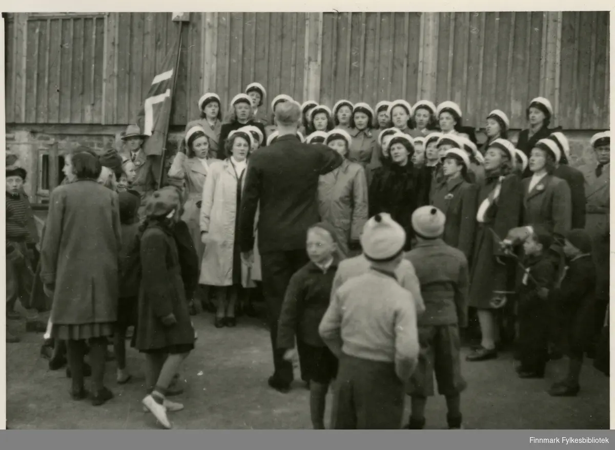 Vadsø damekor synger. Bildet er tatt 17.mai 1949. Ved siden av damene ser man et stort norskeflagg. Kvinnene har på seg hvite hatter (trolig kor uniform) og kåper. Det er barn blant tilskuerne. Jentene har på seg skjørt og lange jakker. Jenten under flagget har på seg sko, sokker og lue. Guttene ved siden av henne har på seg shorts og knebukser. En mann ser ut til å dirigere koret. Ved siden av norskeflagget står en til mann, han har på seg hatt.