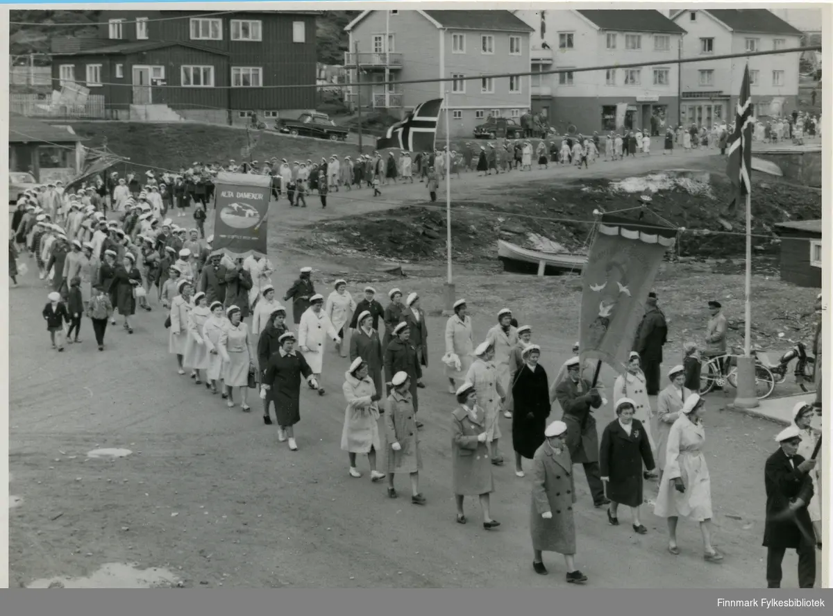 Bildet er tatt i Berlevåg 1962 og viser trolig et 17.mai tog. Flere av personene på bildet har på seg en hvit hatt, trolig kor uniform. Avbildet på bildet er flere av Vadsø damekors medlemmer, og enkelte mannskor medlemmer. I toget kan man se at det bæres faner og ved siden av veien er det satt opp norskeflagg. I bakgrunnen kan man se hus, parkert utenfor husene kan man se biler. Ved siden av den høyre flaggstangen kan man se en sykkel, ved siden av den trolig en moped.