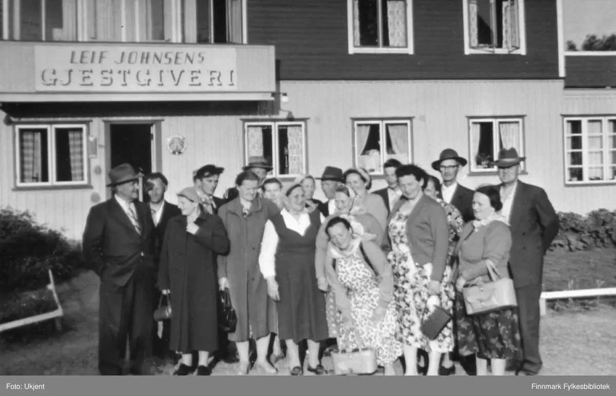 Nesseby husmorslag er ute å reiser. Her poserer de foran Leif Johnsens Gjestgiveri, Rustefjelbma, Tana. Kvinnene har på seg kjoler, skjørt, skjorter, strømpebukser og sko. Enkelte har på seg skaut, andre har briller. Kvinnene holder på vesker. På veggen kan man se vinduer med gardiner i.Blant kvinnen kan man se enkelte menn. Mennene har på seg hatter, jakker, slips og bukser.