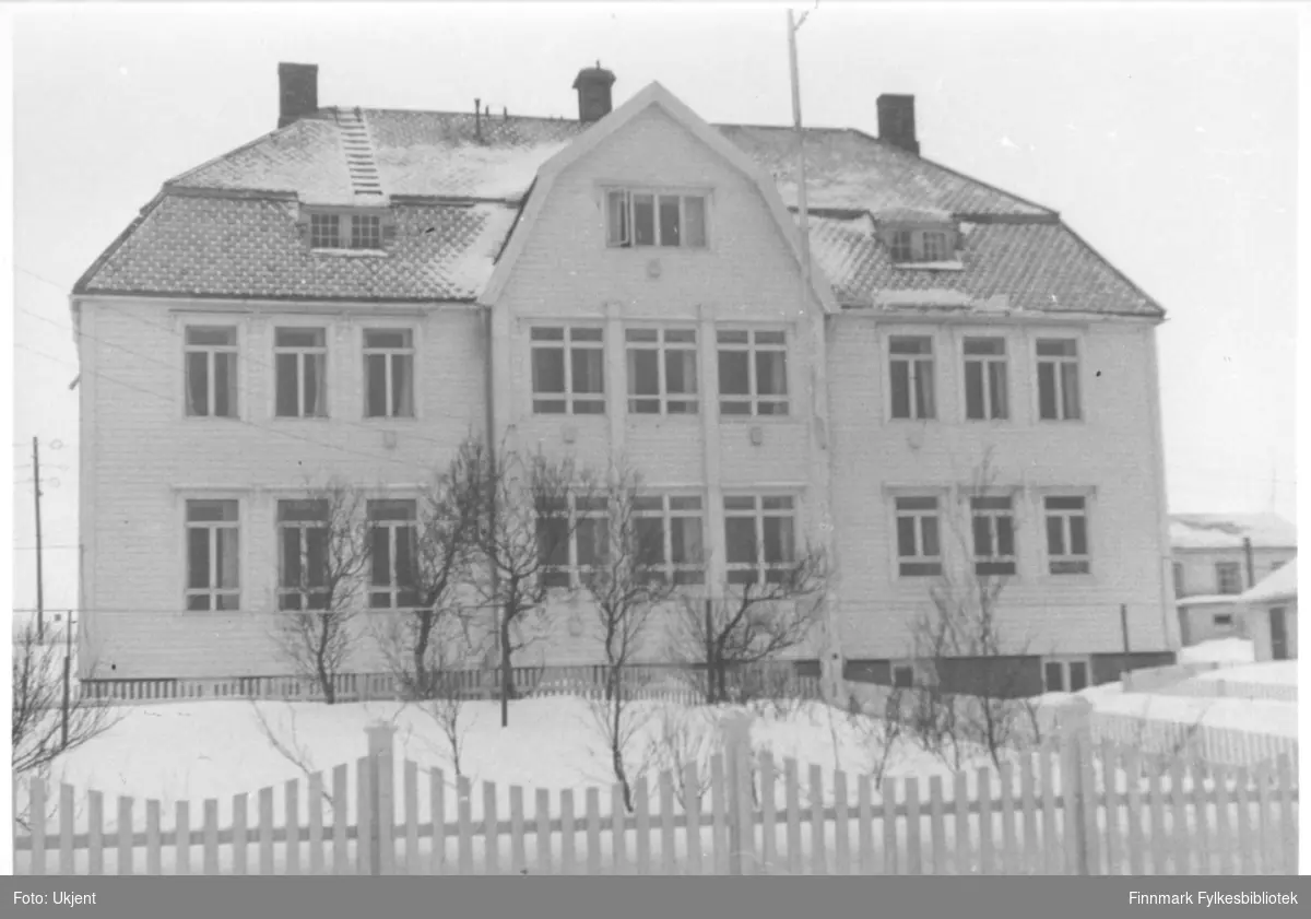 Vadsø sykehus på 1950-tallet. På bygget kan man se vinduer, skorsteiner og en grunnmur. På taket kan man også se en stige som fører opp. Foran Sykehuset kan man se et lukket området omringet av gjerder. I dette området vokser det også trær. Bak sykehuset kan man skimte et annet bygg.