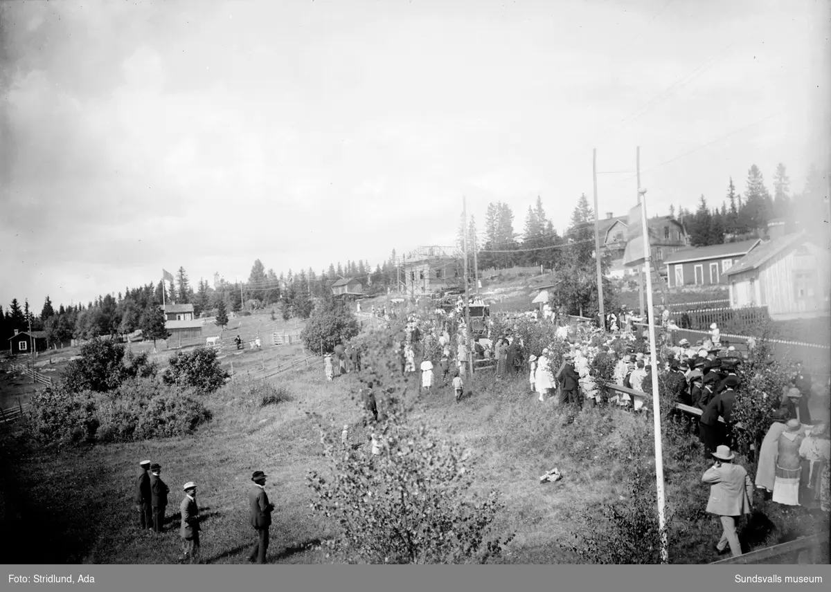 Vy. "Barnens dag på Alnö"  Vägen (rondellen och norrut) är nuvarande Alnövägen, och i bildens underkant byggdes senare nerfarten till Alnö Folkets park (kallat "Burken"). Huset längst till höger där åkaren och dragspelaren Harald Karlsson slog sig ned med familj. Uthuset bakom är känt som kioskägaren Teodor och Olga Hamrins. Olga född Hamberg. Hamrins var verksamma till omkr. 1950. Lite till vänster bakom uthuset syns Axel Carlssons fastighet (samtida med Hamrins), samt ännu högre upp ses tak med skorsten, tillhörande Wii skola, som brann omkr. 1940. Mitt i bilden ett pågående husbygge av och för Henning Hambergs småbarnsfamilj.