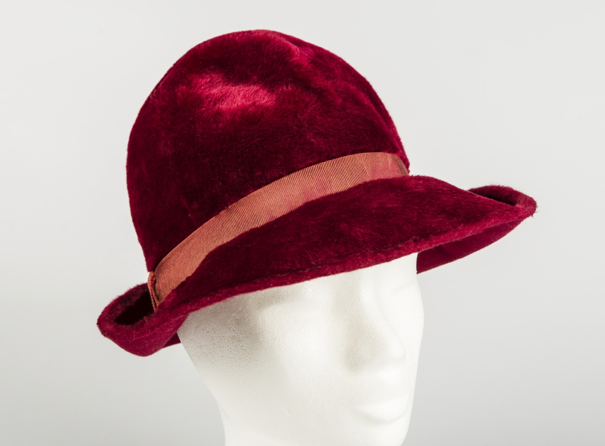 Håndsydd, burgunderrød fedora med høy, rund pull, nestet på innsiden, slik at den får to bulker på siden foran à la fedora herrehatter. Også toppen av den høye, runde pullen, er nestet på innsiden slik at den trekkes ned i en bulk. Relativt lang silkefloss. Smalt ripsbånd omkring pullen, med dekorativt element der båndet møtes bak på høyre side. Relativt bred brem, mykt oppoverbøyd og med smal falding på undersiden. Innvendig svettebånd av ripsbånd.