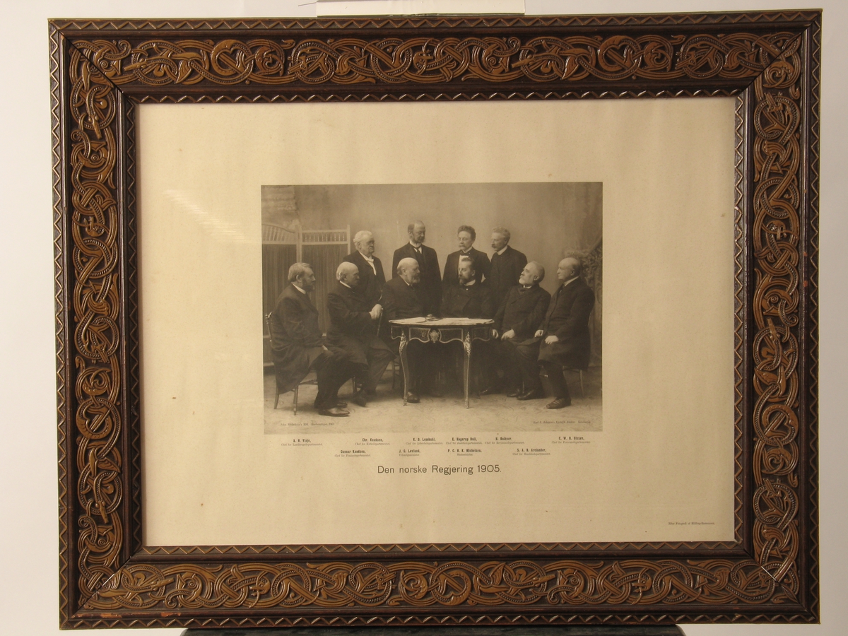 Gruppeportrett av den norske regjering i 1905,  10 menn samlet rundt et bord. 6 sitter på stoler, 4 stående bak. De 5 på høyre side ser mot venstre, og vise versa. Navnene er trykt i bildets nedkant.