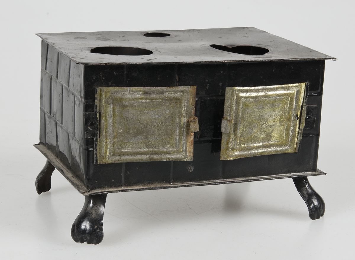 Leksaksspis av svartmålad plåt med två luckor på framsidan, löstagbar skorsten, två hål på ovansidan för kastruller samt fyra fötter. Till spisen hör tre kastruller av plåt, omålade. Två med gulfärgat lock och en utan lock med långt handtag.