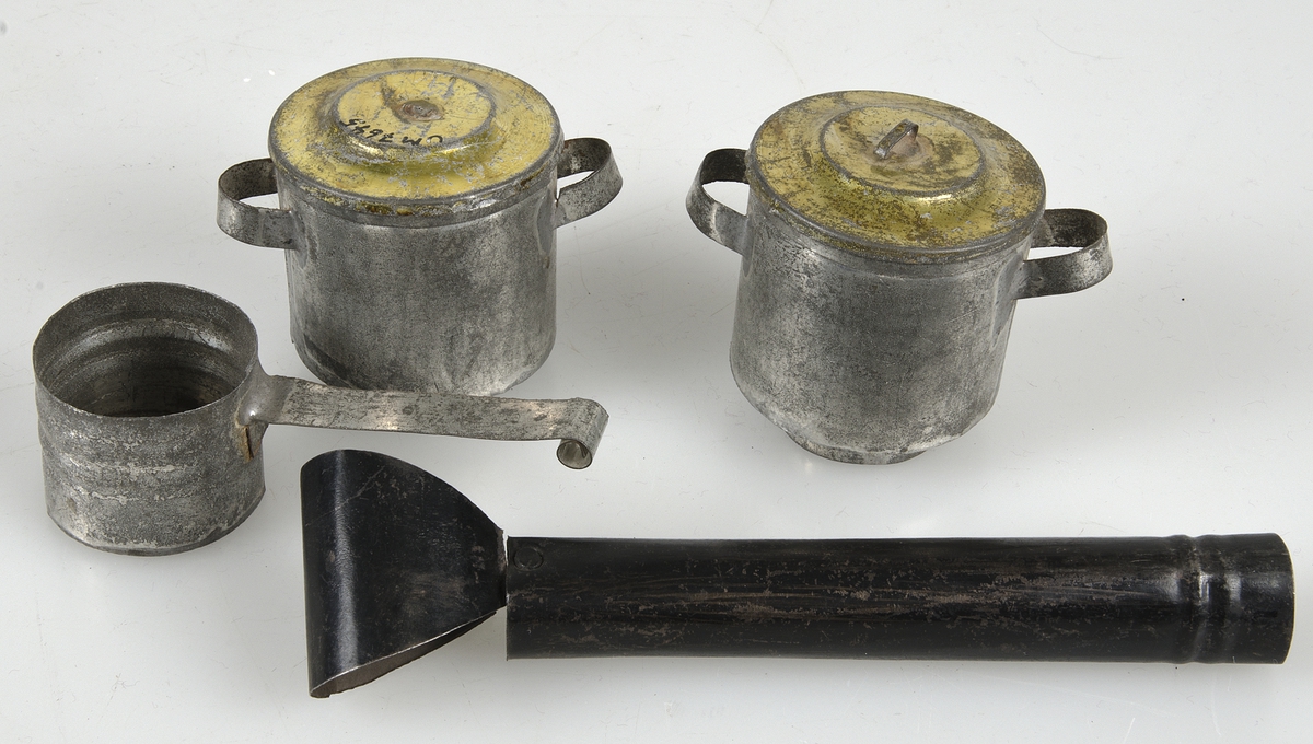 Leksaksspis av svartmålad plåt med två luckor på framsidan, löstagbar skorsten, två hål på ovansidan för kastruller samt fyra fötter. Till spisen hör tre kastruller av plåt, omålade. Två med gulfärgat lock och en utan lock med långt handtag.