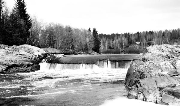 Fra det øvre Flisfallet i elva Flisa i Åsnes kommune i Hedmark. Fotografiet er tatt fra østre elvebredd, i motstrøms retning, mot en terskel der elva ser ut til å ha et fall på halvannen til to meter. I bakgrunnen, på østre side av elva, skimter vi et par bygninger.