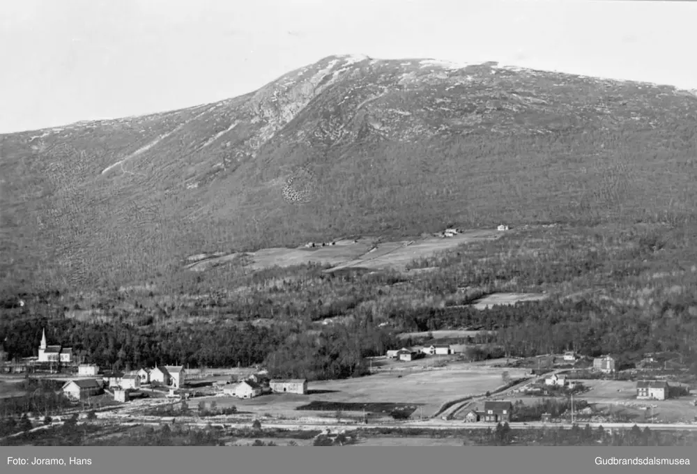 Oversiktsbilde av ulike bygninger og natur rundt Lesjaskog stasjon, Raumabanen, fjell og li i bakgrunnen.