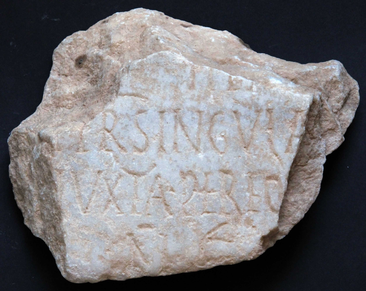 Fragment av marmortavle  med antikk innskrift. Gulhvit I  grovkornet   marmor.  Innskrift hvorav leselig  Tilst. 1972 god. Bruddflatene og oversiden noe mer skitne  (værslitte?) enn undersiden(baksiden), som har fals for o  ppfesting langs nedre del. 
