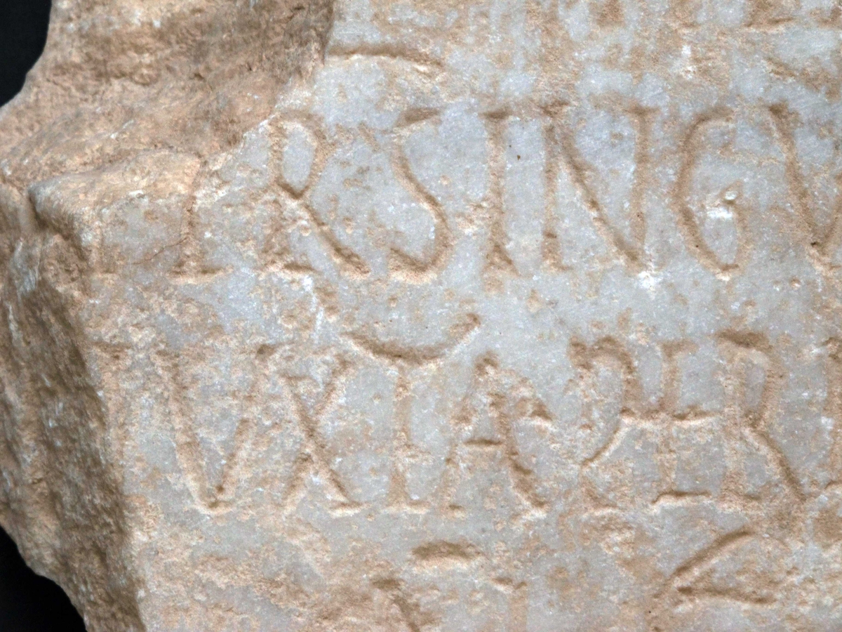 Fragment av marmortavle  med antikk innskrift. Gulhvit I  grovkornet   marmor.  Innskrift hvorav leselig  Tilst. 1972 god. Bruddflatene og oversiden noe mer skitne  (værslitte?) enn undersiden(baksiden), som har fals for o  ppfesting langs nedre del. 

