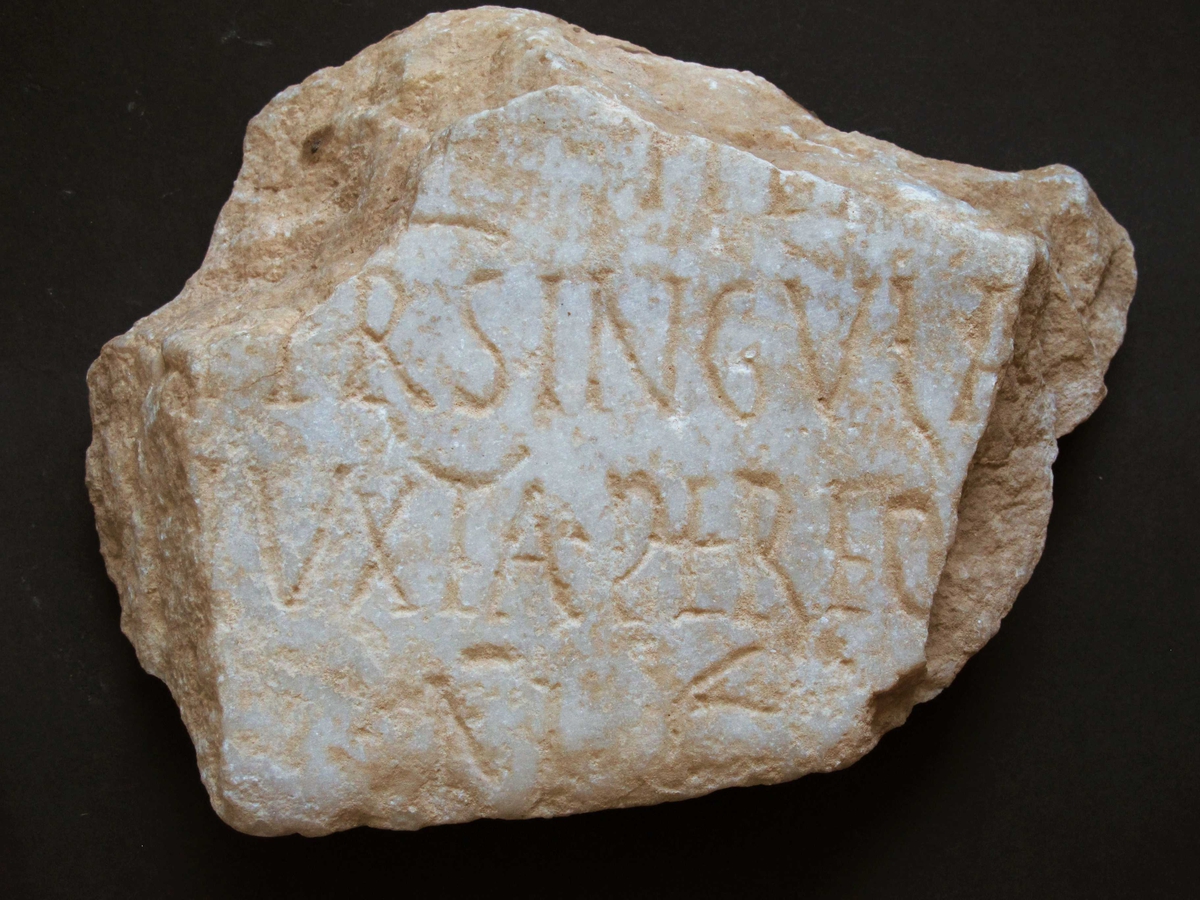 Fragment av marmortavle  med antikk innskrift. Gulhvit I  grovkornet   marmor.  Innskrift hvorav leselig  Tilst. 1972 god. Bruddflatene og oversiden noe mer skitne  (værslitte?) enn undersiden(baksiden), som har fals for o  ppfesting langs nedre del. 
