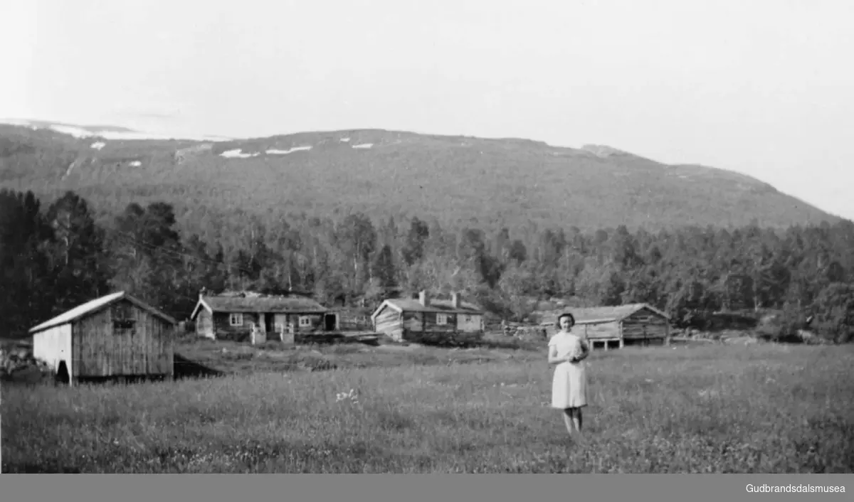 Kvinne står ute på jordet på Øygardssetra, det er fire bygninger bak, til venstre er Sør-Skotte, til høyre Sør-Slettom.