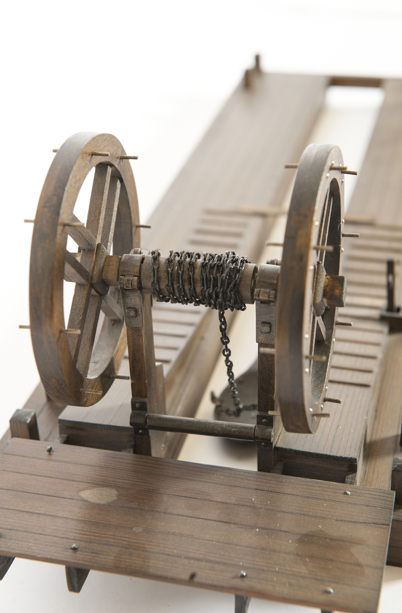 Modell av furu, betsad. Flotte, tudelad, jämte mudderskopa och liggande spel. Dubbla pollare i alla hörn samt två pumpar på flotten.