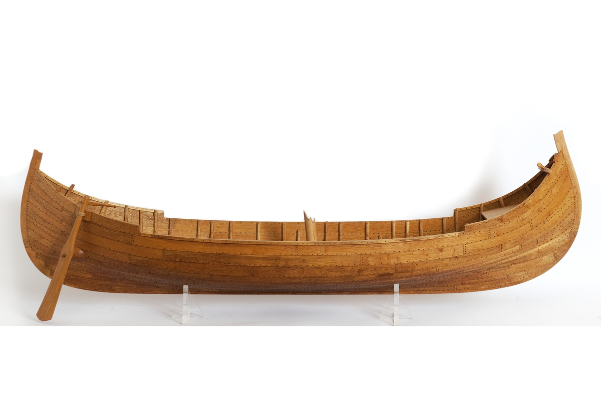 Fartygsmodell av ek, klinkbyggd på spant, högbordigt skepp (knarr)  ÄSKEKÄRRSBÅTEN  (fynd år 1933 fr. Göta älv, daterat 700 e. Kr.) Sidoroder på styrbordssidan, bordläggning förhöjd med ett bord i för- och akterskepp, liten back och halvdäck, kort kölsvin med mastspår.