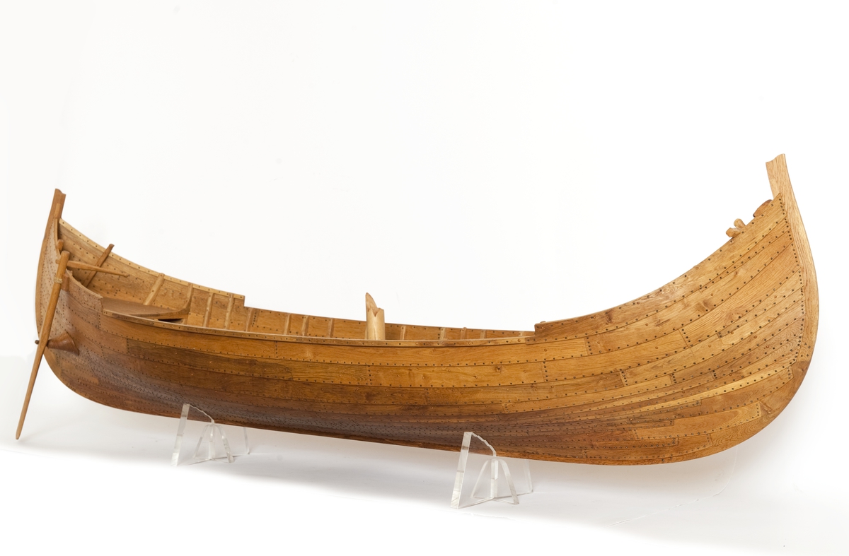 Fartygsmodell av ek, klinkbyggd på spant, högbordigt skepp (knarr)  ÄSKEKÄRRSBÅTEN  (fynd år 1933 fr. Göta älv, daterat 700 e. Kr.) Sidoroder på styrbordssidan, bordläggning förhöjd med ett bord i för- och akterskepp, liten back och halvdäck, kort kölsvin med mastspår.