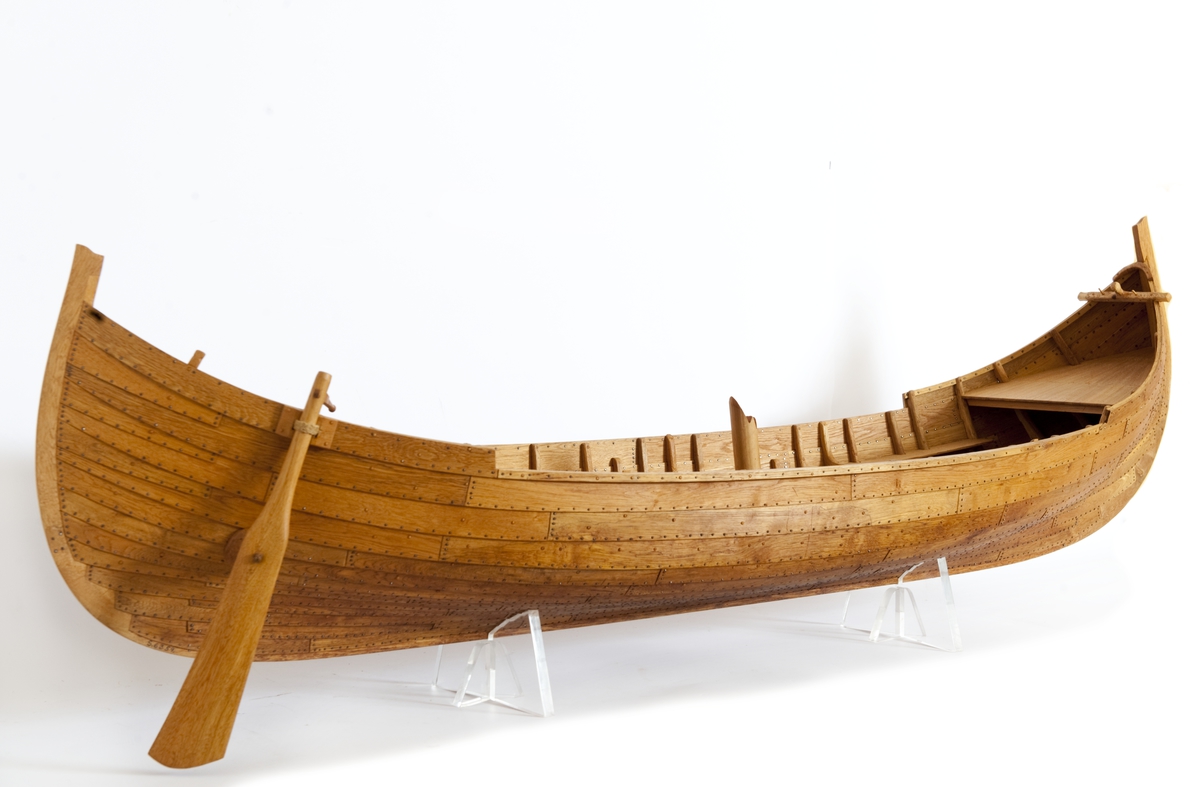 Fartygsmodell av ek, klinkbyggd på spant, högbordigt skepp (knarr)  ÄSKEKÄRRSBÅTEN  (fynd år 1933 fr. Göta älv, daterat 700 e. Kr.) Sidoroder på styrbordssidan, bordläggning förhöjd med ett bord i för- och akterskepp, liten back och halvdäck, kort kölsvin med mastspår.