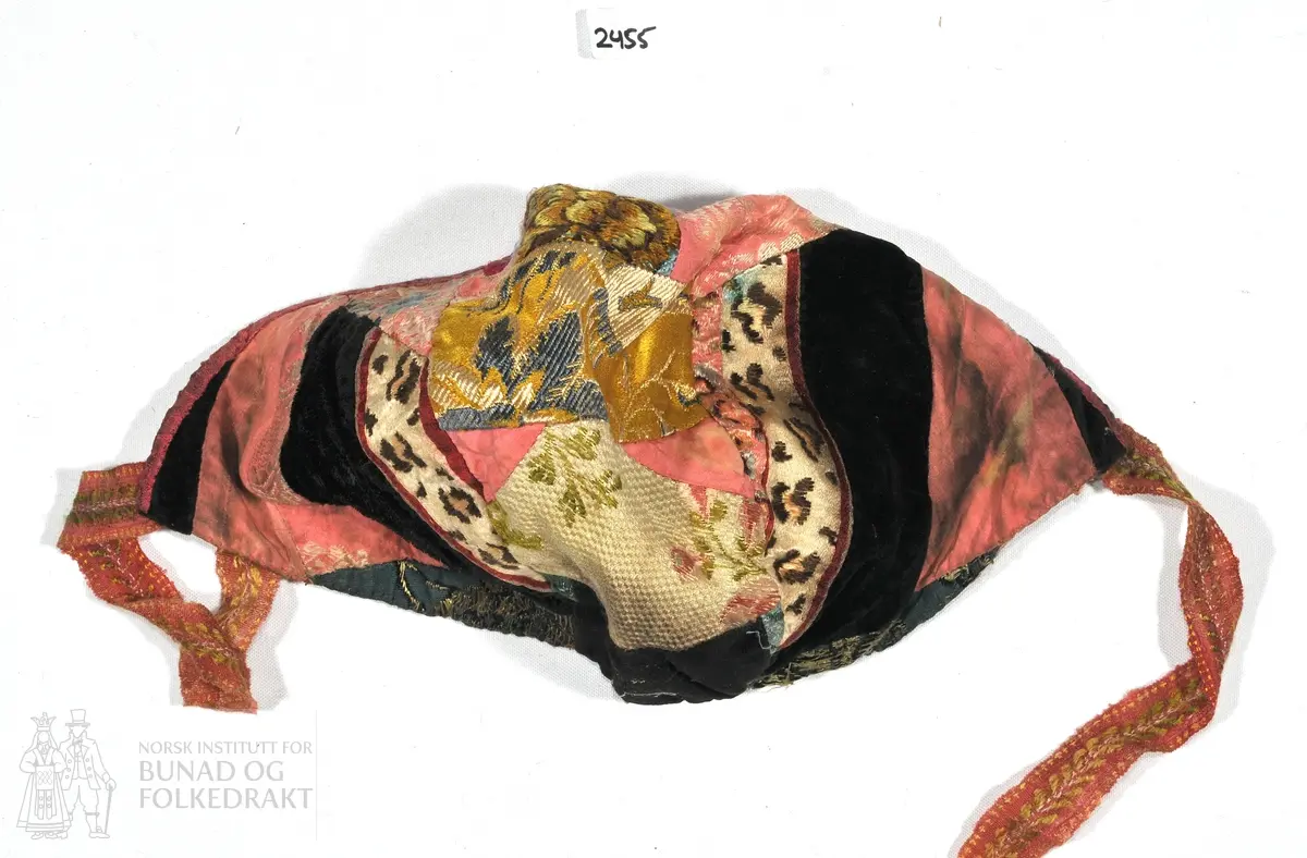 Barnelue sydd sammen av flere ulike stoffbiter av silke og fløyel. Linlerret i foret, kantet med rosa smalt bånd i nakken. Mønstret knyteband. Lengde over pannen; 28cm. lengde over nakken; 28cm.