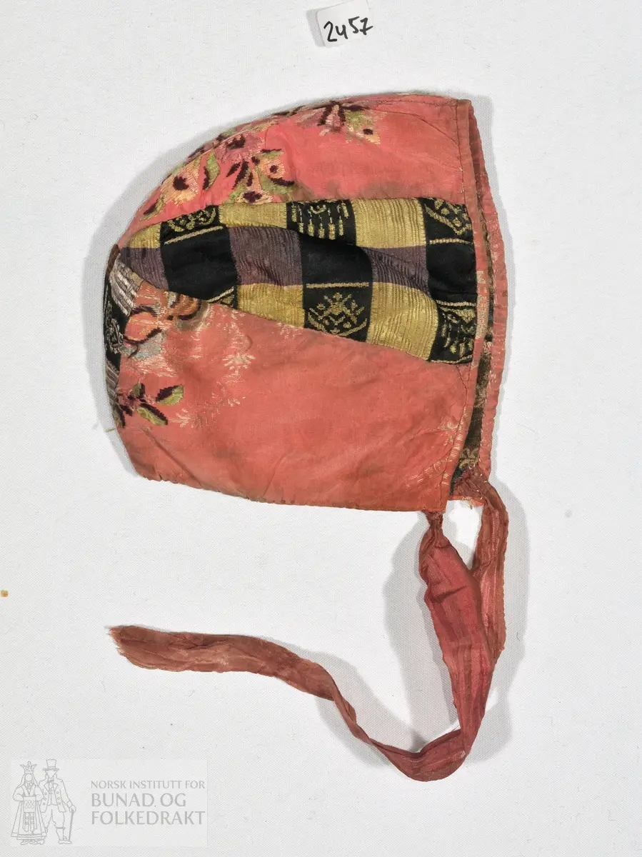 Dåpslue i rosa brosjert silkedamask, og rutet silke.  Stripet trykt, bomullsstoff i for. er kantet med et rosa silkestoff av samme kvalitet som i kilene.
Sydd av seks trekanter, guttelue.

Lengde fra øre til øre:
1. over nakken: 24cm.
2. over panna: 27cm.