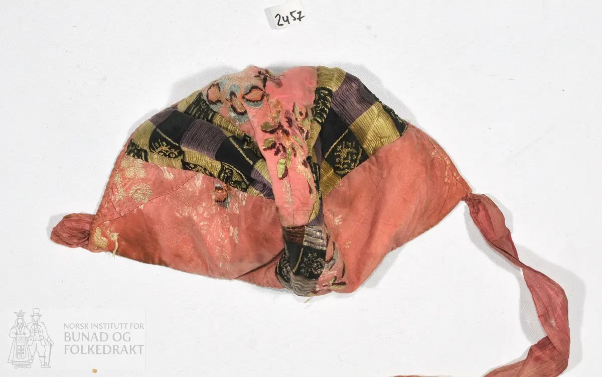 Dåpslue i rosa brosjert silkedamask, og rutet silke.  Stripet trykt, bomullsstoff i for. er kantet med et rosa silkestoff av samme kvalitet som i kilene.
Sydd av seks trekanter, guttelue.

Lengde fra øre til øre:
1. over nakken: 24cm.
2. over panna: 27cm.