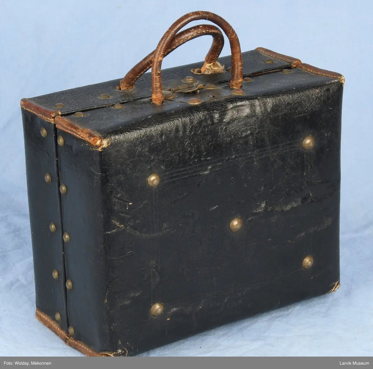 Form: Liten koffert med lås, buet håndtak på hver del. Inni er den delt i to med en plate som kan spennes fast med to stropper. Dekorert med metall kn
