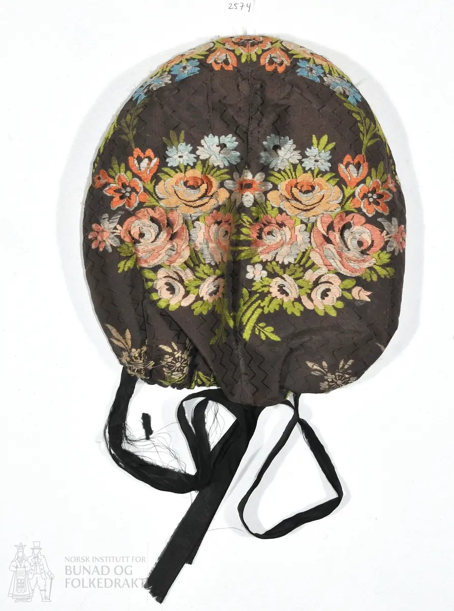 Botthuve i brosjert silke. Pull satt sammen av to stoffestykker. Vattert oppe og midt bak, men ikke nede ved rynkingen i nakken. Kantet med rosa silkeånd og foret med trykt, glansa bomull. Håndsøm. 

Lengde fra øre til øre over panna: 40cm.