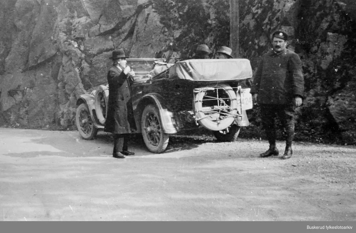 Rutebileier Adolf Karlsen står parkert langs veien med sin bil av typen Minerva, skiltnummer F2424, ved Skaret.
En mann står og røyker utenfor bilen, imens to passasjerer sitter i baksetet. Antageligvis er det Adolf Karlsen som står til høyre i bildet.