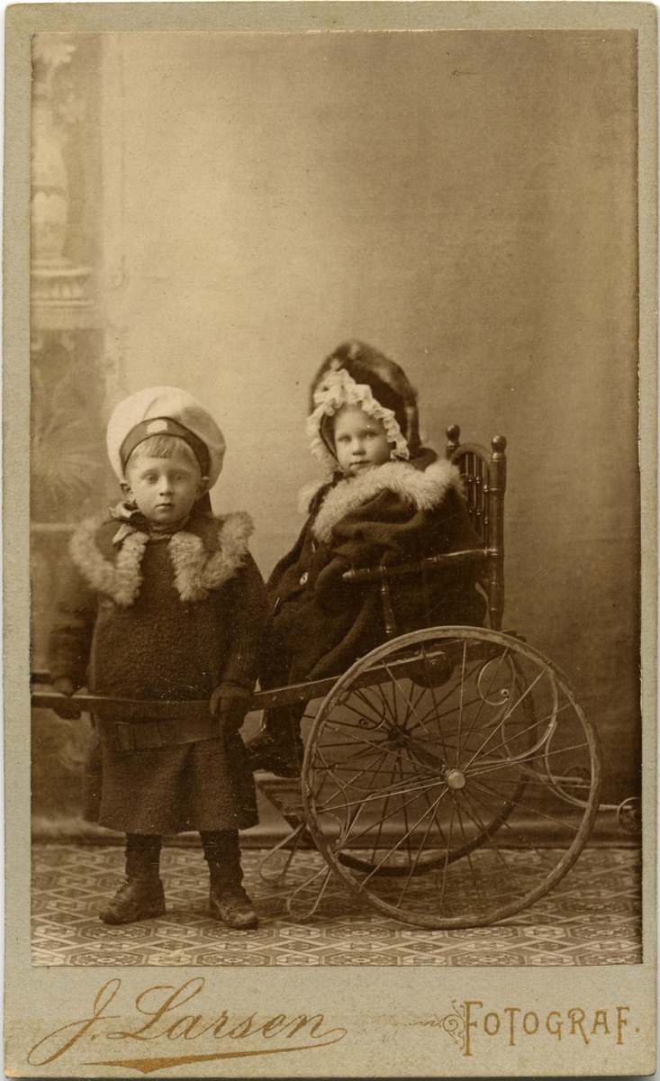 To ukjente barn poserer for et bilde. En liten jente sitter i en trekkbar stol og en gutt står ved siden av.