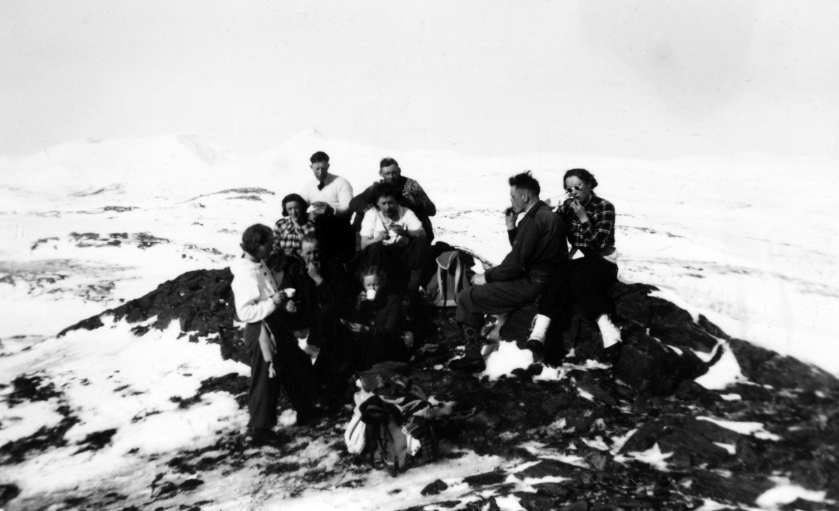 Menn og kvinner har kaffepause på en fjelltopp. Snødekte fjell i bakgrunnen.