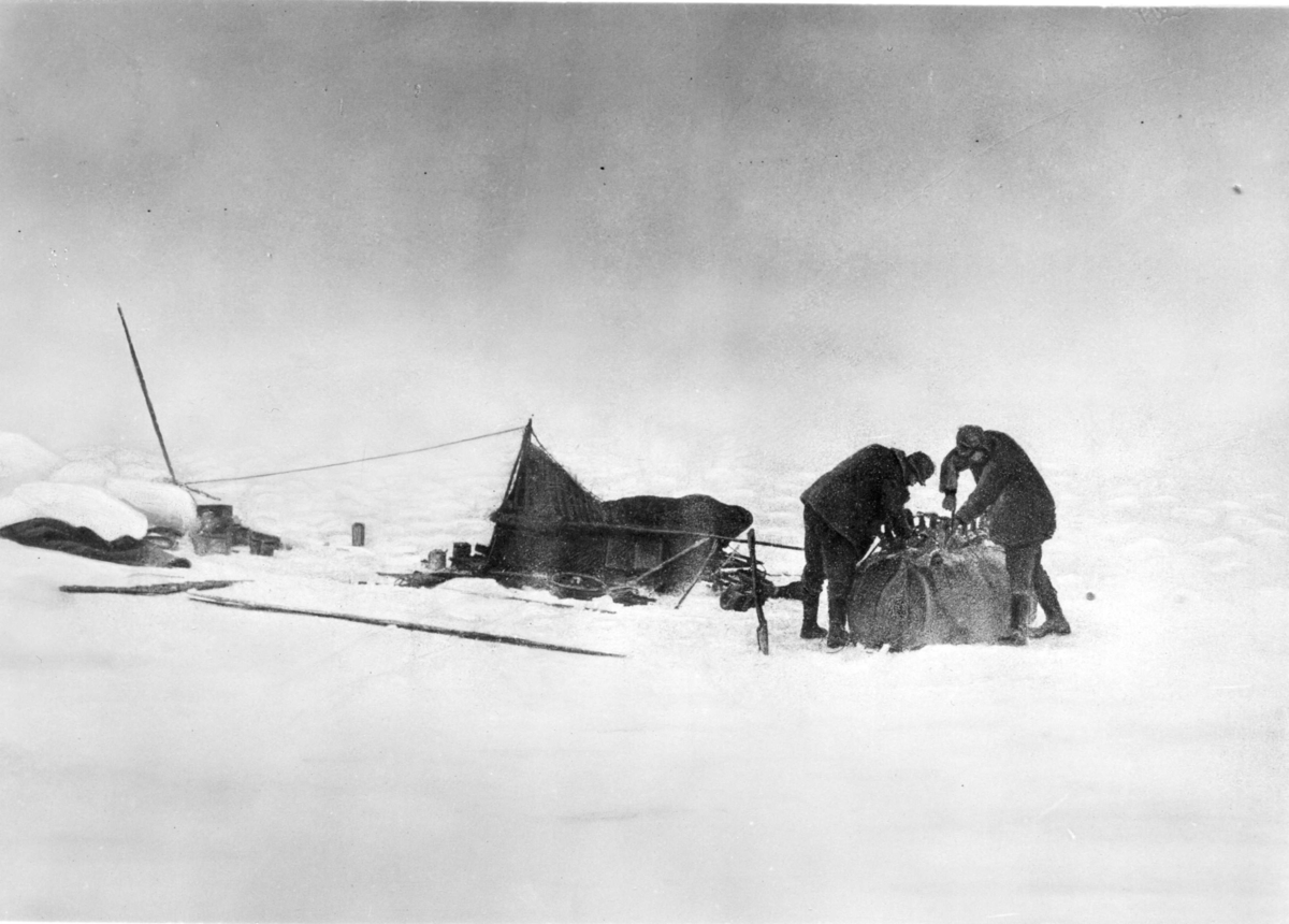 Isvandringen. S A Andrée till vänster och Knut Fraenkel arbetar på släden som är surrad vid den upp och nedvända båten. Tälten till vänster. Retuscherad variant av gm.III-82.