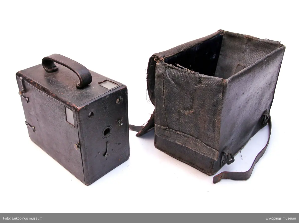 Lådkamera med väska, väskan av duk. Insidan trä klädd med svart sammet. Remmar av läder. Kameralådan av trä klädd med läder. Handtag på ovansidan av läder. Detaljer av metall och glas. Serienummer 3100 på båda kortsidorna. Mitt på ena kortsidan inpräglat märke som dock endast går att tyda- EXPRESS.