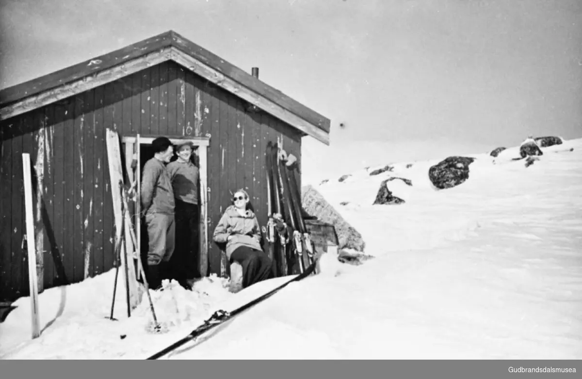 Vangsbue ved Hårrtjørnet, tre personer i husveggen, det er vinter, de er ute på ski, ski og staver står oppreist langs veggen. 