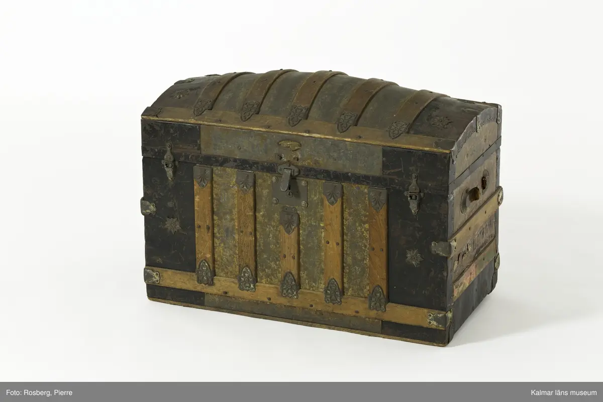KLM 28210. Koffert, trä, metall, papp. Kista av trä med metallbeslag, välvt lock, lås, två spännen och beslag i form av sköldar med blommor. Delvis svartmålad med spår av gul färg. Handtag har suttit på gavlarna, båda saknas. Kofferten har haft fyra hjul av troligtvis mässing på underredet, endast ett av dessa är kvar. Invändigt klädd med papp med småblommigt mönster i grönt på gul botten. På lockets insida två fack. det högra facket har ett papper klistrat i botten med mönster i orange, blått och gult på grön botten och blommig bård i blått och grönt. På höger gavel påskrift: ? LSTRÖM ? SWEDEN.